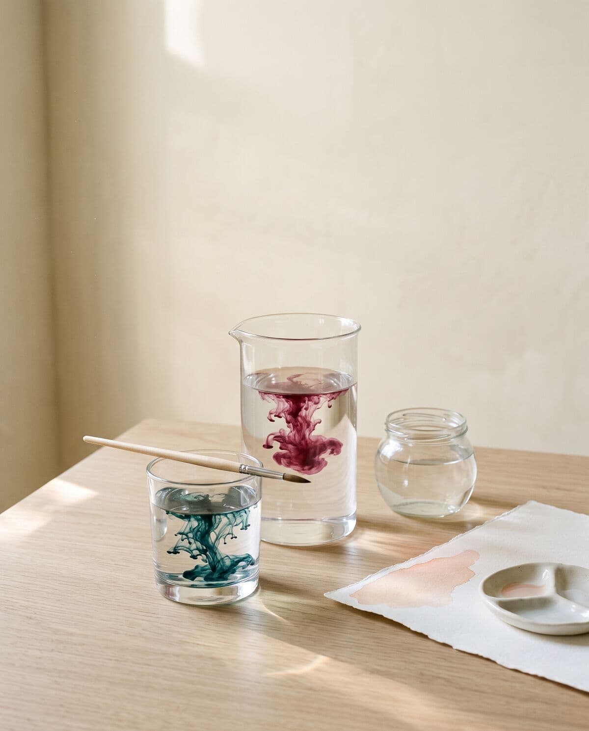 Vasos de vidrio con agua sobre una mesa de roble: en uno se abre una gota de pigmento magenta y en otro una azul petróleo; al lado, un pincel, papel de acuarela con una aguada durazno y una paleta de cerámica