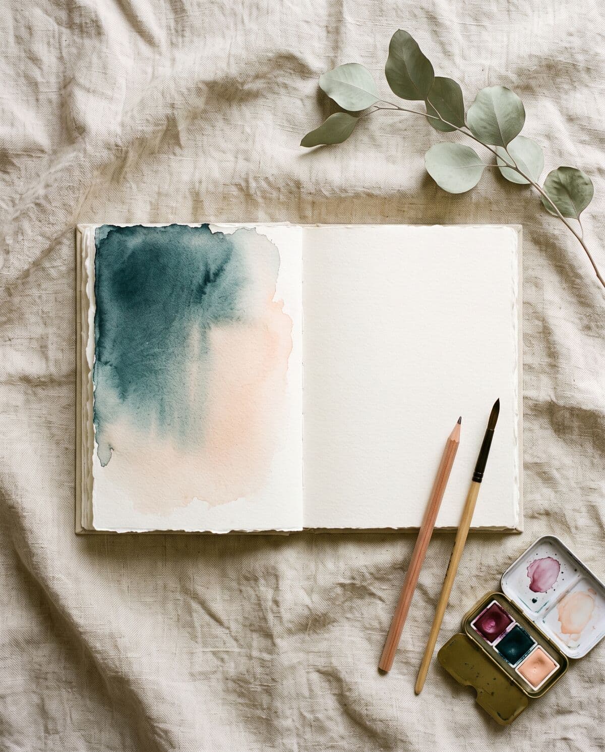 Cuaderno abierto con una aguada azul petróleo en una página y la otra en blanco, junto a un lápiz y una cajita de acuarelas